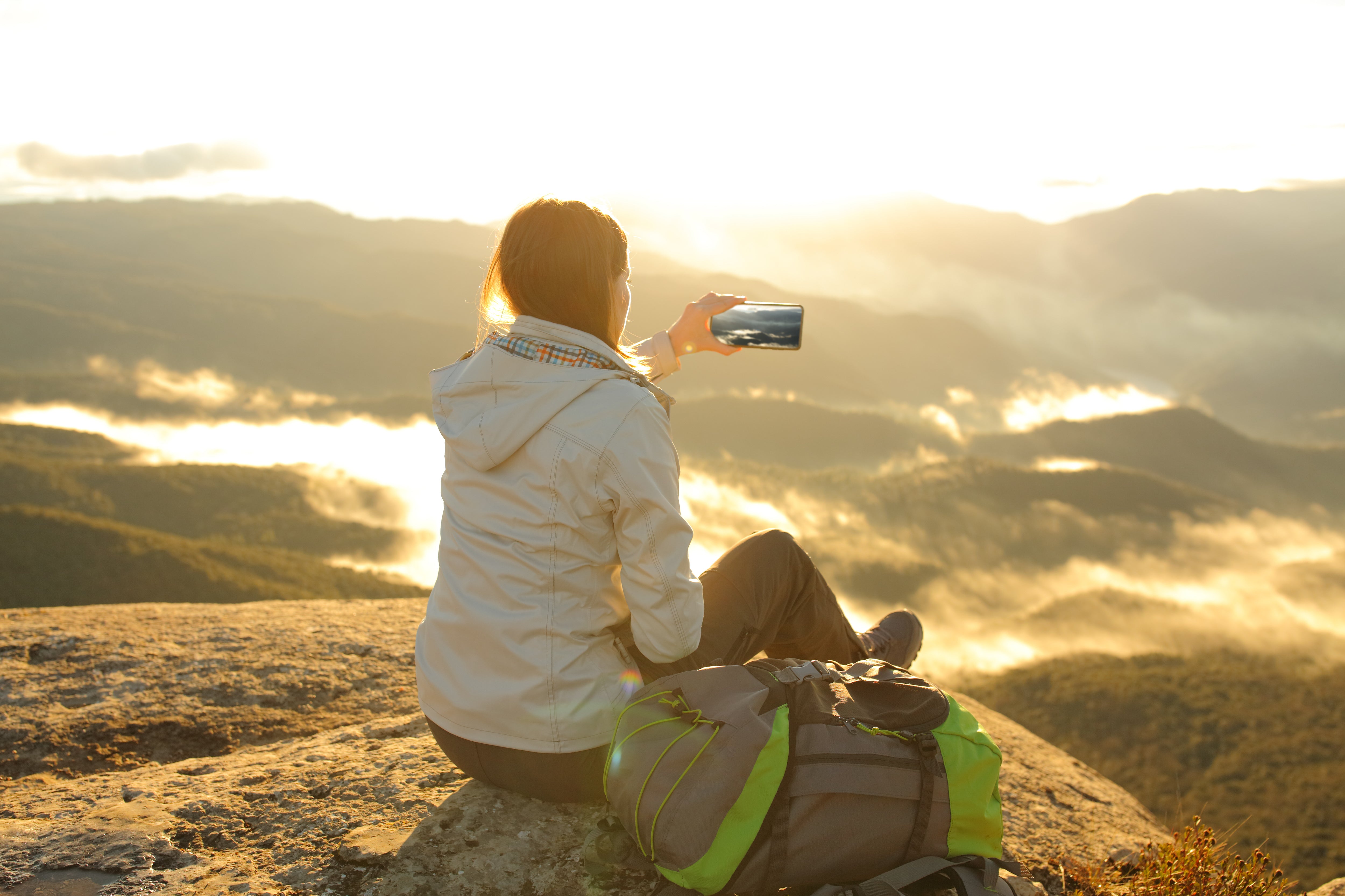 une femme de dos prend une photo avec son téléphone portable, elle capture la montagne, son sac à dos de randonneuse posé à côté d'elle 